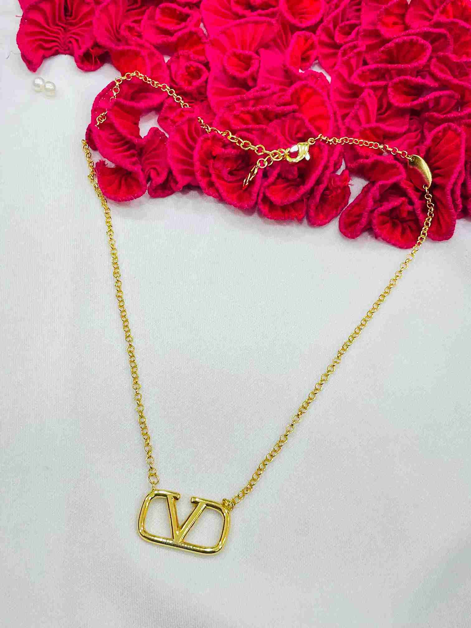 V Classic Gold Chain with bold V logo pendant on a fine gold chain displayed on a white surface with red fabric roses as props