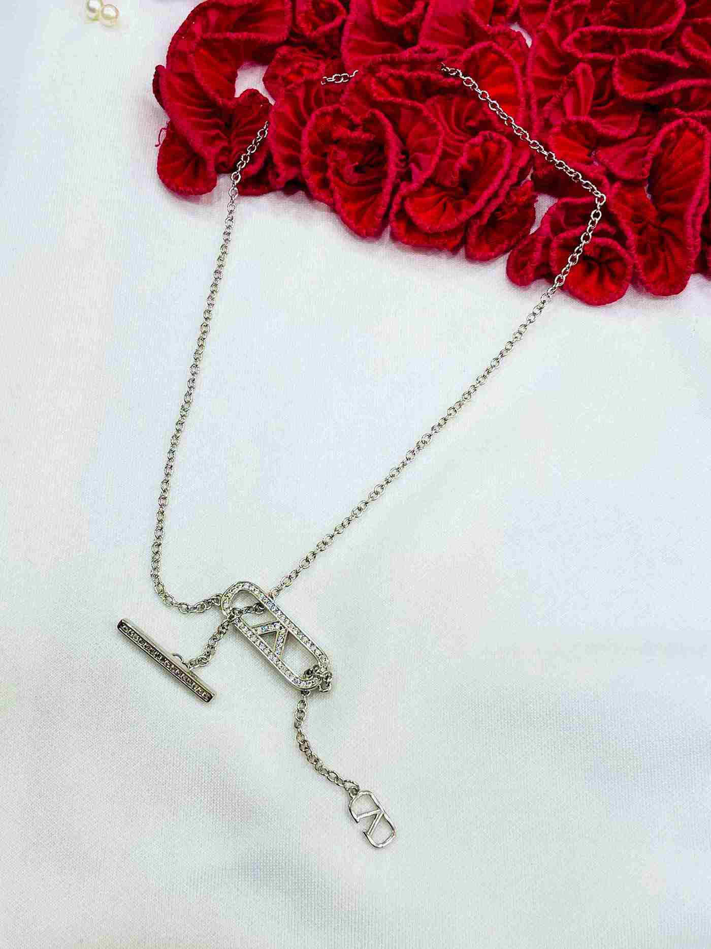 V Bling Chain crystal-studded V logo and bar pendant on a silver chain displayed on white surface with red fabric roses as props